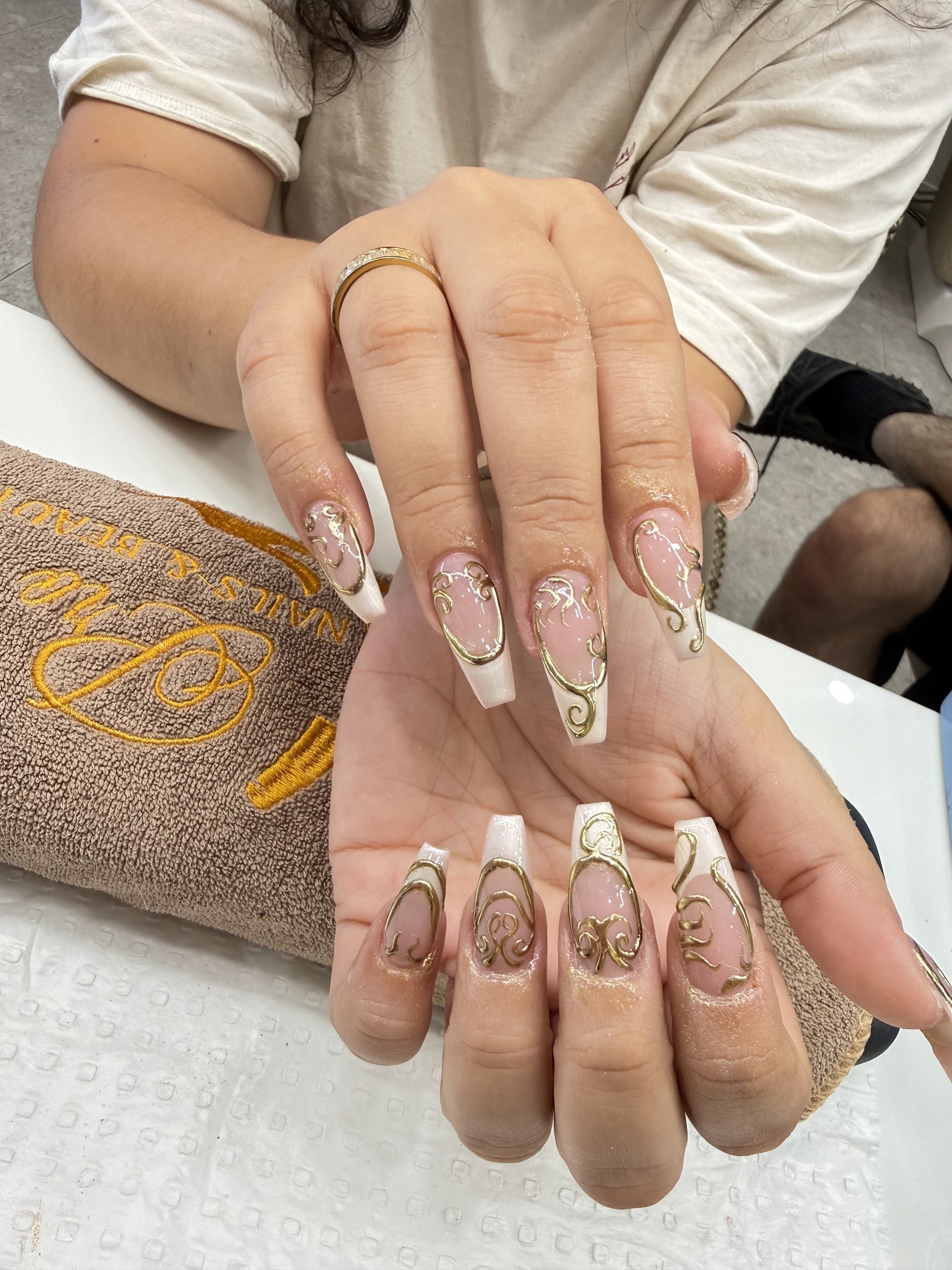 White and clear ombré acrylic nails with fine gold filigree detail, resting on a Pro Nails & Beauty salon towel
